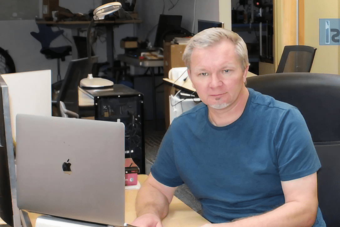 A man sitting at a desk with a laptop in a cluttered room.